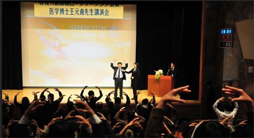 图4：二零一二年一月十四日，王元甫在东京新宿的会堂演讲“不花钱、不吃药的气功健康法”。图为，王元甫在讲台上演示法轮功第二套功法——法轮桩法，台下与会者跟着学炼动作。